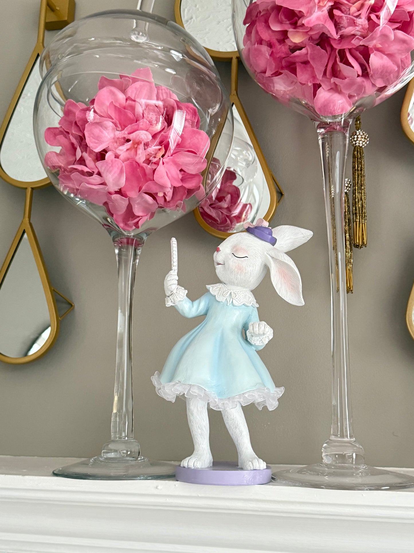 white bunny dressed in a blue dress with white ruffled trim, a purple hat, and holding a small mirror.